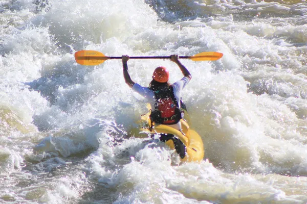 A person in a yellow kayak raising a paddle above their head as a large wave crashes into the front of the kayak.