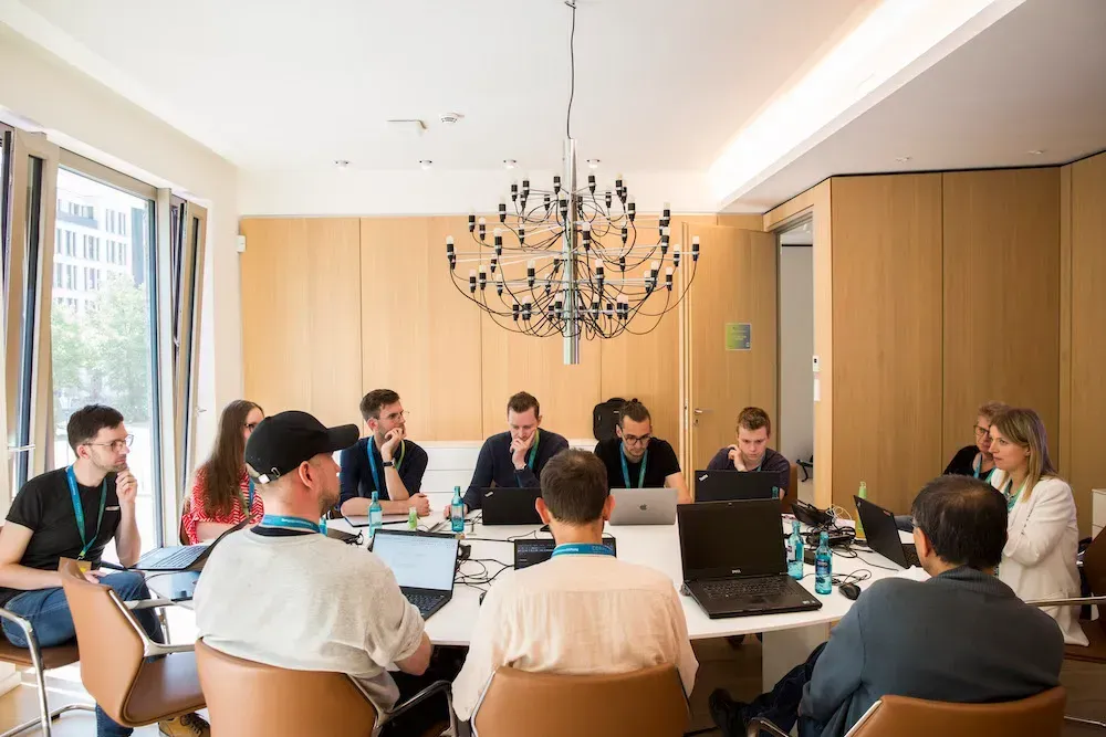 Eine Gruppe von ca 10 Personen sitzt um einen Tisch in einem Konferenzraum. Die meisten haben einen Laptop vor sich.