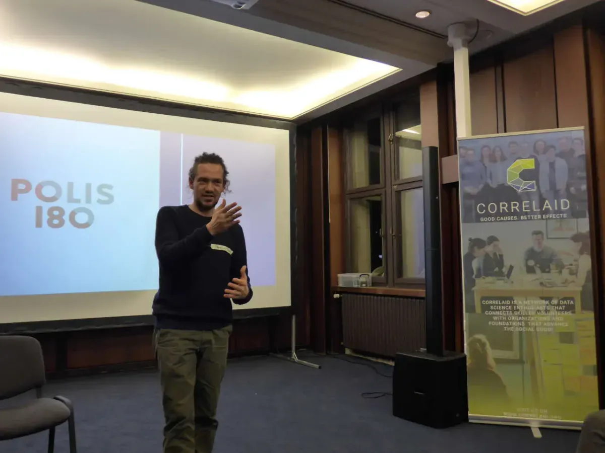 A person stands in a room and recites something. She gestures while doing so. In the background, POLIS 180 can be read on a screen. On the right of the picture is a roll-up advertising CorrelAid.