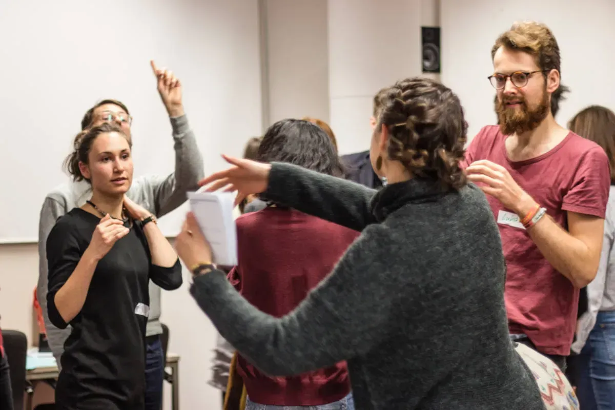 Mehrere Menschen stehen einander zugewandt. Zwei Personen schauen auf eine dritte, die gestikuliert. Im Hintergrund meldet sich eine Person.