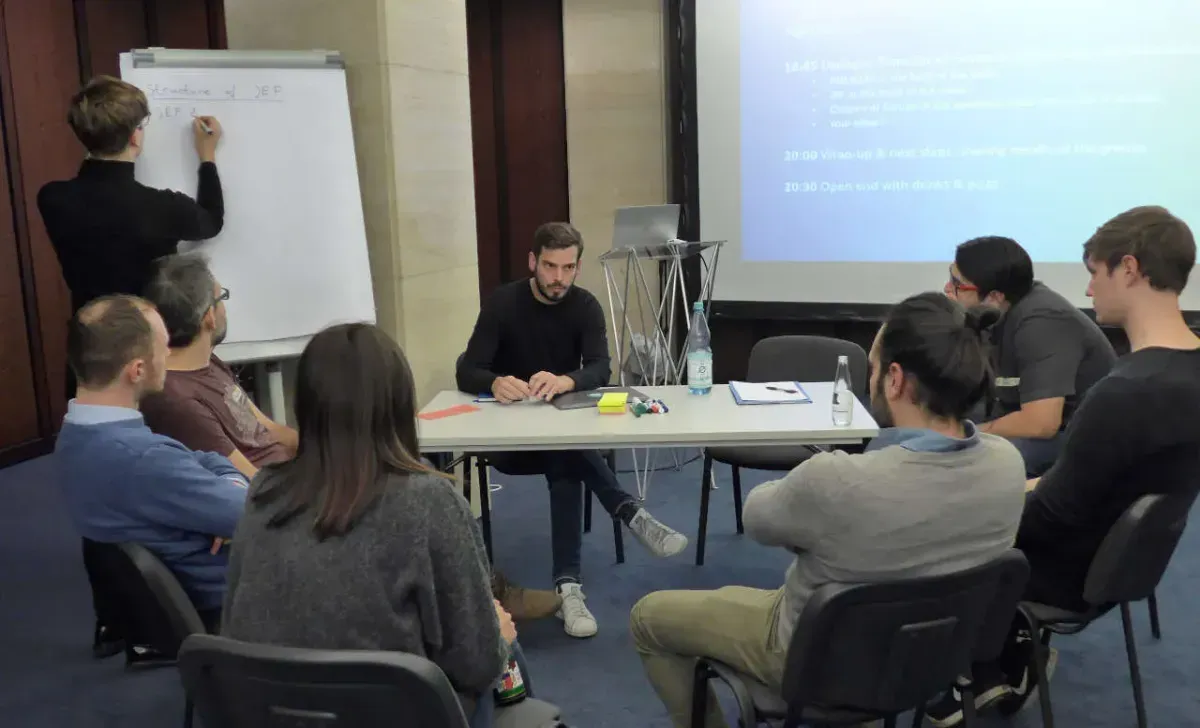 a group of people working together and discussing. One person is sitting at a white table with 6 people sitting in a semicircle around the table. One person in the background is standing and taking notes on a flipchart