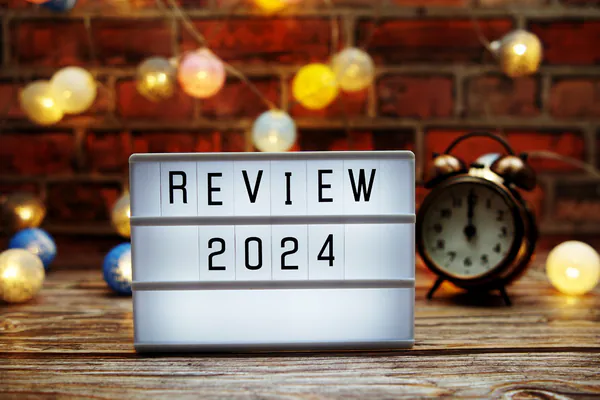 Foto einer Leuchttafel mit der Aufschrift: Review 2024, im Hintergrund sind kleine Licher und einen Wecker der Mitternacht zeigt