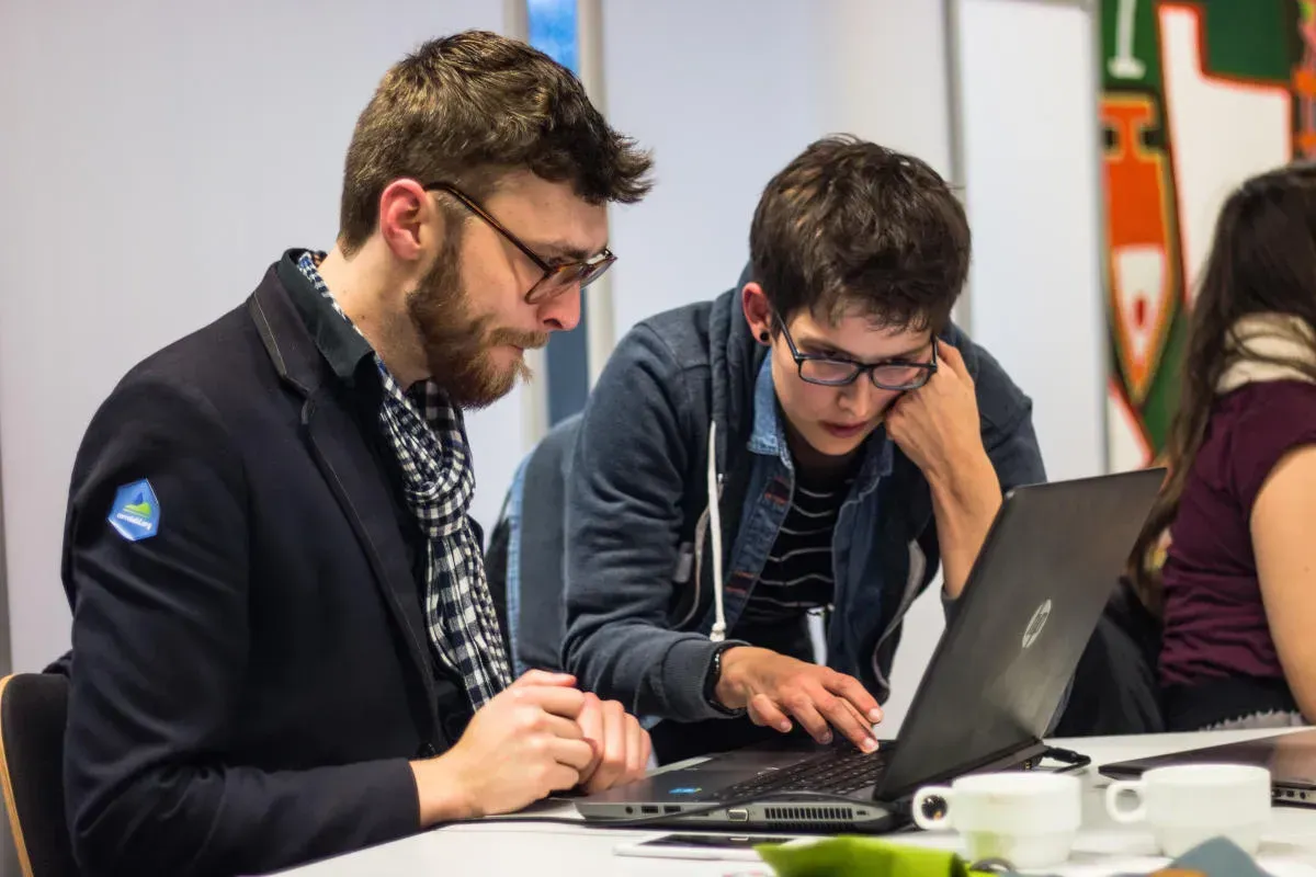Eine Person sitz vor einem Laptop und schaut mit einer andern Person zusammen auf den Bildschirm. Die andere Person steht gebeugt. Beide sehen konzentriert aus.