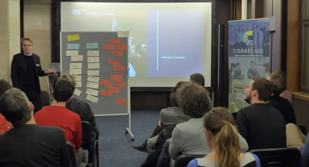 A person is presenting a number of post-its that are pinned on a whiteboard. A group of approximately 10 people is seated in rows of chairs and is listening attentively. In the background, there is a roll-up advertising CorrelAid.