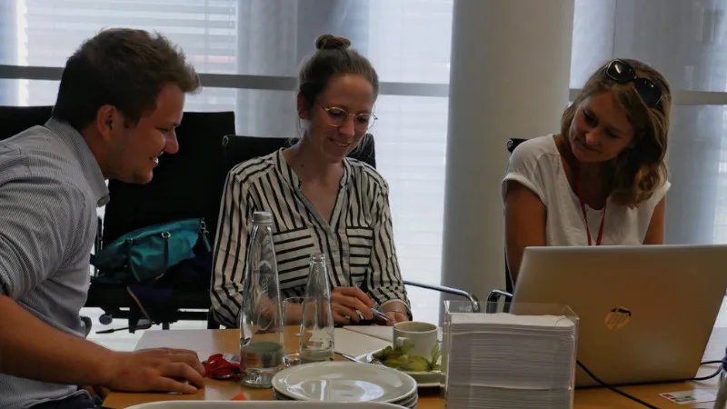 Three people sit at a table. On the table there is an open laptop, two bottles, a plate and a coffee cup. The three people are smiling to each other