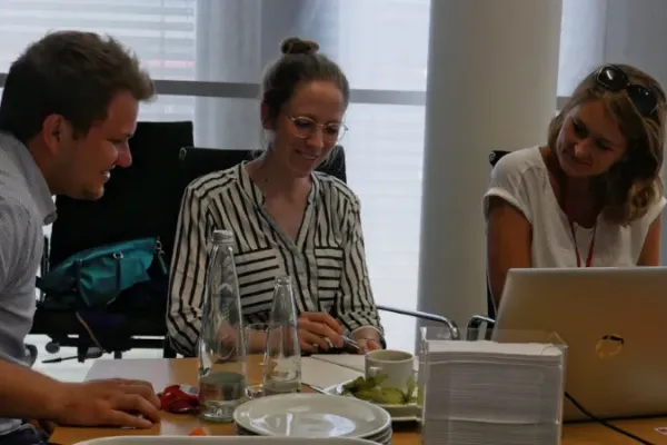 Three people sit at a table. On the table there is an open laptop, two bottles, a plate and a coffee cup. The three people are smiling to each other