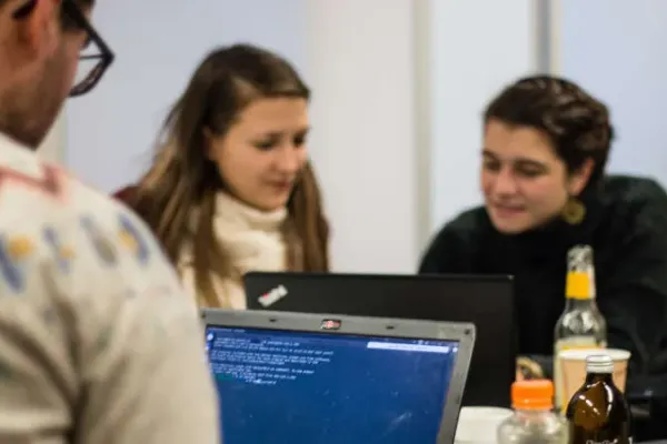 Man sieht den Rücken und den Kopf einer Person, die am Laptop sitzt. Zwei Personen im Hintergrund sitzen zusammen an einem Laptop und unterhalten sich angeregt. Auf dem Tisch stehen verschiedene Flaschen, z.B. Fritzlimo.
