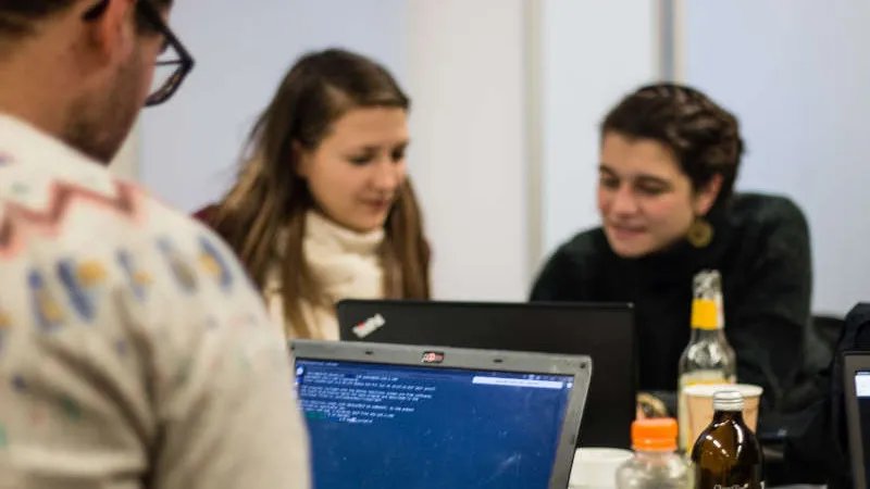 Man sieht den Rücken und den Kopf einer Person, die am Laptop sitzt. Zwei Personen im Hintergrund sitzen zusammen an einem Laptop und unterhalten sich angeregt. Auf dem Tisch stehen verschiedene Flaschen, z.B. Fritzlimo.