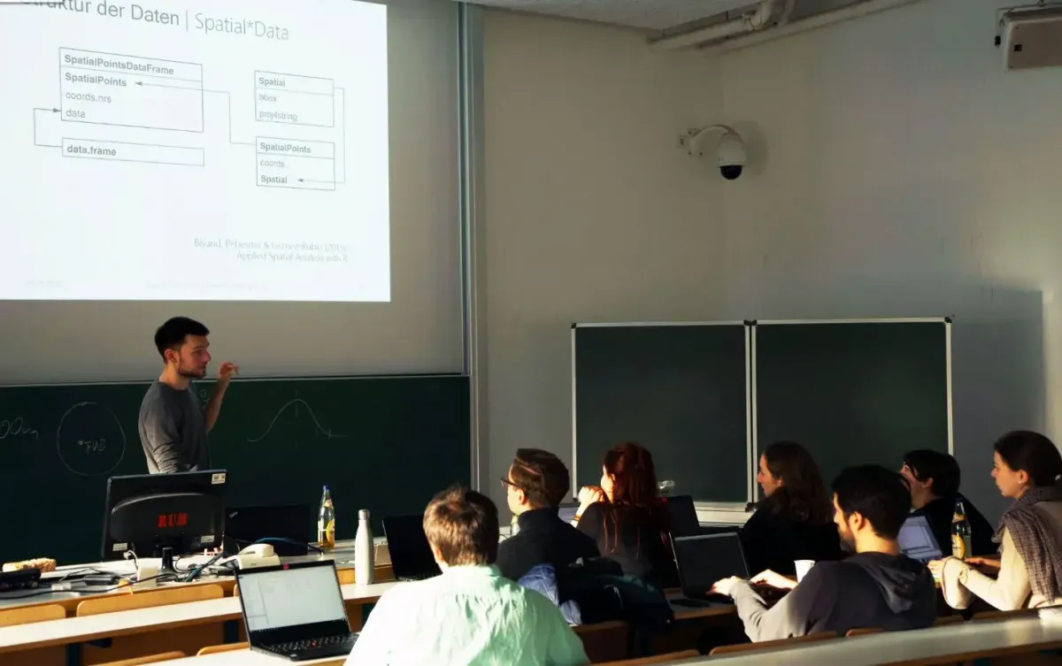 Bild eines Seminarraums. Eine Person steht vorne und erkl&auml;rt einer Gruppe von ca. 10 sitzenden Personen etwas. Im Hintergrund die an die Leinwand geworfenen Folien. Alle Personen haben einen Laptop vor sich. 