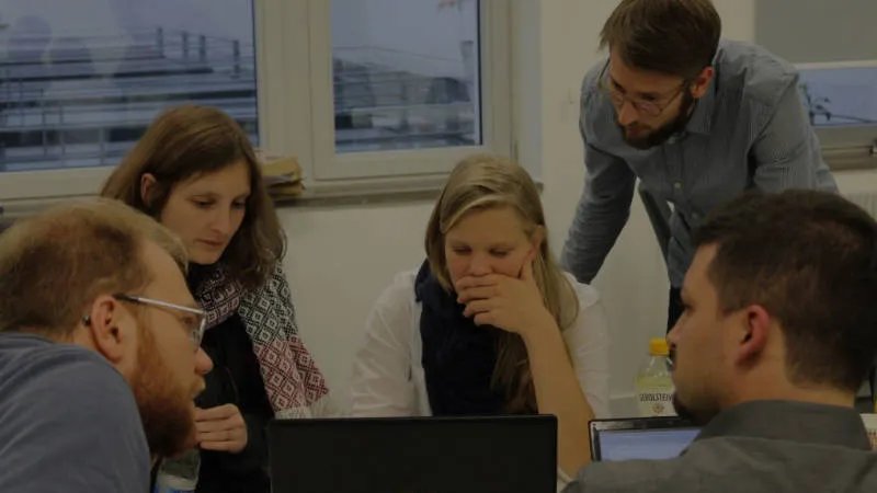 eine Gruppe von Personen zwischen 20 und 30 Jahren sitzt an einem Tisch und schaut auf einen Laptop.