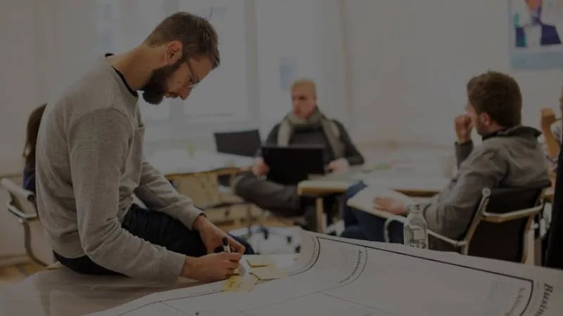 Eine Person mit braunen Haaren und Vollbart sitzt auf einem Tisch und schreibt auf Post-It Karten. im Hintergrund sitzen zwei weitere Personen und unterhalten sich. Eine Person hat einen Laptop auf dem Schoß. 