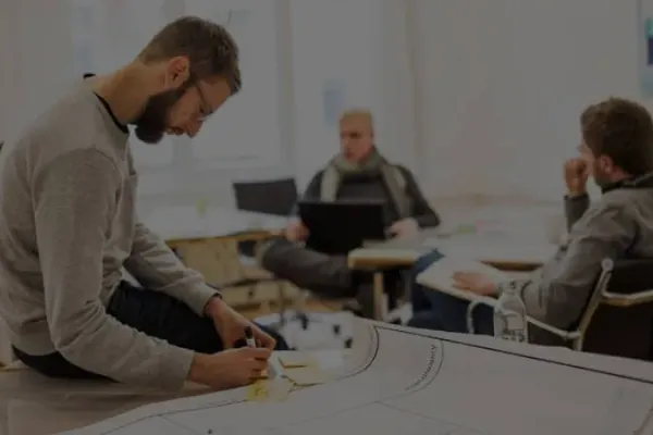 Eine Person mit braunen Haaren und Vollbart sitzt auf einem Tisch und schreibt auf Post-It Karten. im Hintergrund sitzen zwei weitere Personen und unterhalten sich. Eine Person hat einen Laptop auf dem Schoß. 