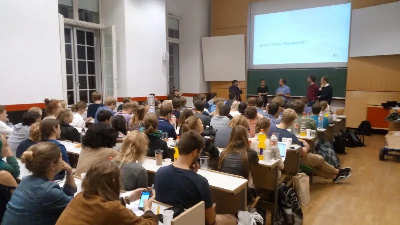 Bild eines Hörsaals, in dem ca. 40 Personen sitzen. Vorne stehen Johannes und vier andere Personen. Auf der Folie, die an die Wand geworfen ist, steht "print("hello Mannheim")