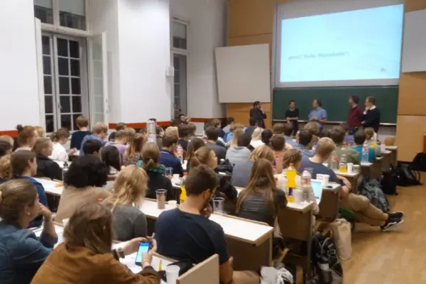 Bild eines Hörsaals, in dem ca. 40 Personen sitzen. Vorne stehen Johannes und vier andere Personen. Auf der Folie, die an die Wand geworfen ist, steht "print("hello Mannheim")