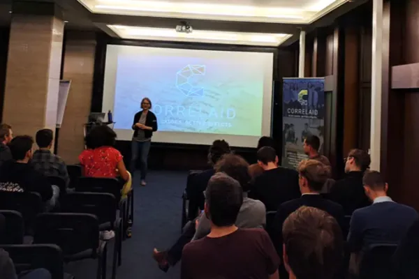Photo of a room with approximately 20 people seated in rows. In front of the rows of chairs is Sylvi who is presenting. Behind her on a canvas is the slide she's currently presenting. It shows the CorrelAid logo against a green/blue gradient background.