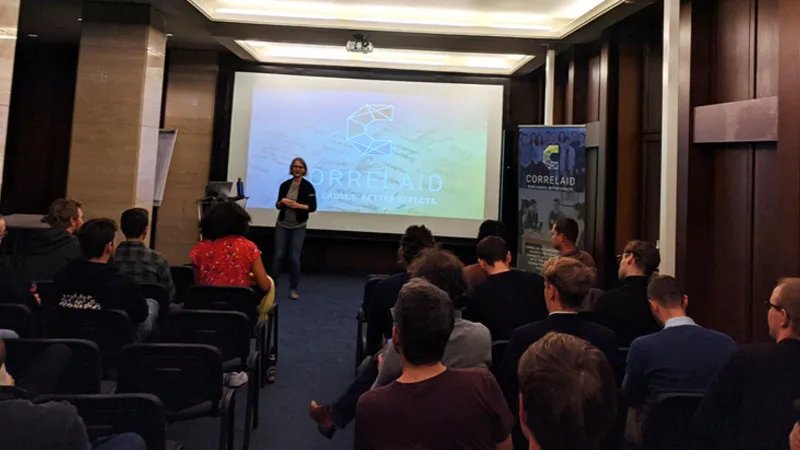 Photo of a room with approximately 20 people seated in rows. In front of the rows of chairs is Sylvi who is presenting. Behind her on a canvas is the slide she's currently presenting. It shows the CorrelAid logo against a green/blue gradient background.