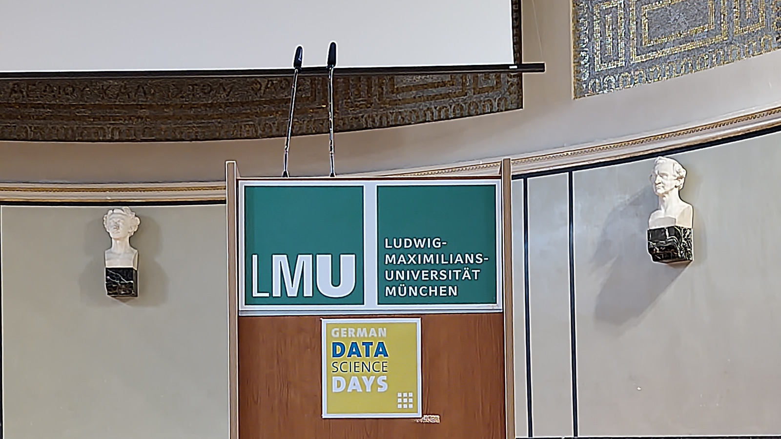 Lectern with LMU and German Data Science Days signage, flanked by two classical busts on the auditorium wall.
