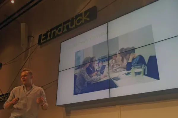 Johannes Müller hält einen Vortrag. Auf dem großen Bildschirm im Hintergrund ist die PowerPoint Präsentation auf der CorrelAider an einem Tisch sitzen. 