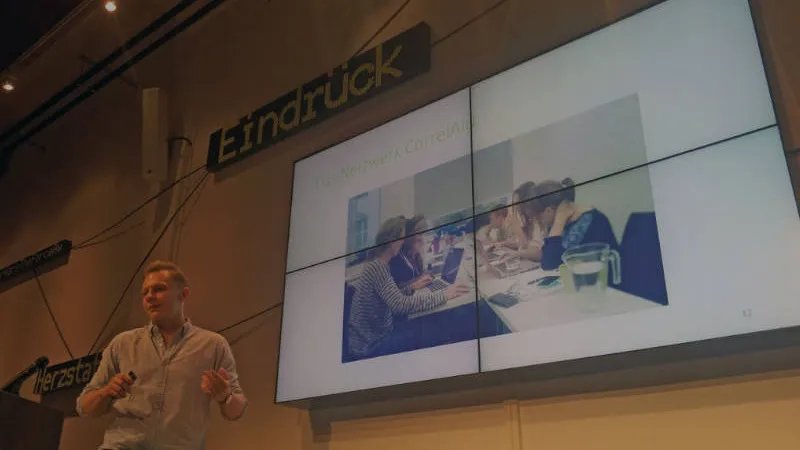 Johannes Müller hält einen Vortrag. Auf dem großen Bildschirm im Hintergrund ist die PowerPoint Präsentation auf der CorrelAider an einem Tisch sitzen. 