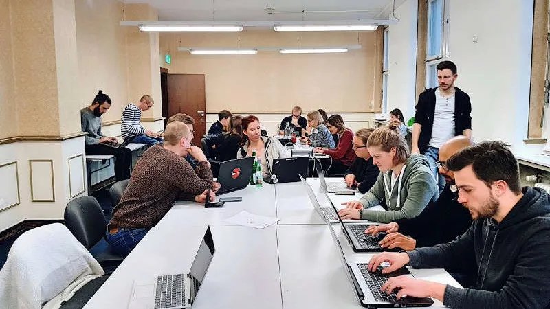 Picture of a seminar room. Approximately 15 people - most of them between 20 and 30 years old - sit around two group tables. They are either working on their laptop or talking to each other. 