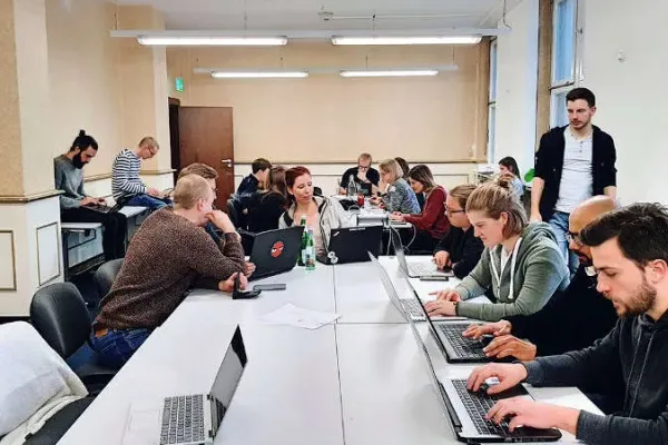 Picture of a seminar room. Approximately 15 people - most of them between 20 and 30 years old - sit around two group tables. They are either working on their laptop or talking to each other. 