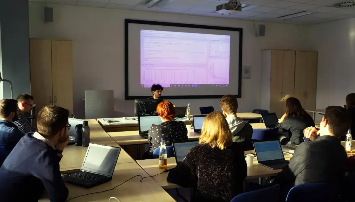 Bild eines Seminarraums in einer Universit&auml;t. Ca. 20 Personen sitzen an Tischen und h&ouml;ren der vortragenden Person zu, die vorne sitzt. Auf der Leinwand hinter dieser Person ist der geteilte Bildschirm der Person zu sehen. RStudio ist ge&ouml;ffnet. Die meisten Personen haben Laptops vor sich. 