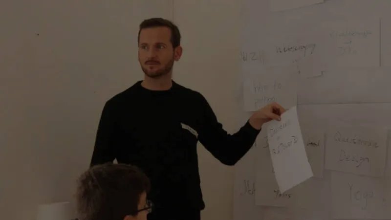 Eine weiße Person mit braunen Haaren, Bart und schwarzem Pullover steht vor einer Wand, an der in großer Schrift beschriebene DinA4 Zettel kleben. Die Person hat einen der Zettel in der Hand und schaut in den Raum hinein.