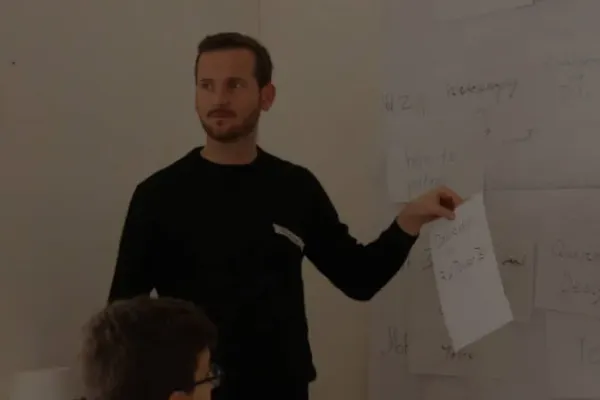 Eine weiße Person mit braunen Haaren, Bart und schwarzem Pullover steht vor einer Wand, an der in großer Schrift beschriebene DinA4 Zettel kleben. Die Person hat einen der Zettel in der Hand und schaut in den Raum hinein.