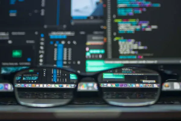 Eine Brille liegt auf dem Trackpad eines aufgeklappten Laptops. Im Hintergrund sieht man verschwommen Code in einer Entwicklungsumgebung auf dem Laptop Monitor.