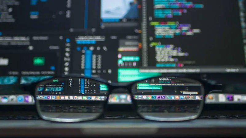 Eine Brille liegt auf dem Trackpad eines aufgeklappten Laptops. Im Hintergrund sieht man verschwommen Code in einer Entwicklungsumgebung auf dem Laptop Monitor.