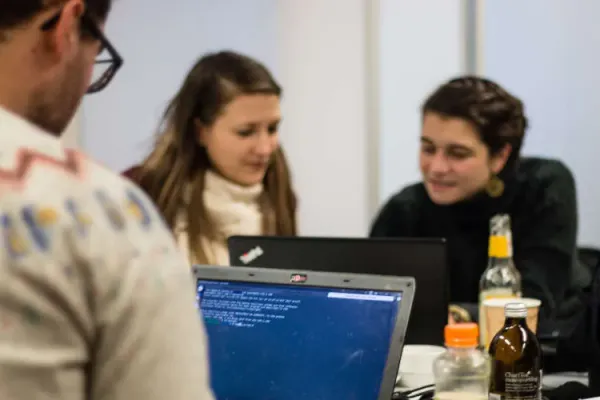 Drei Personen sitzen an ihren Laptops. Zwei von ihnen unterhalten sich.