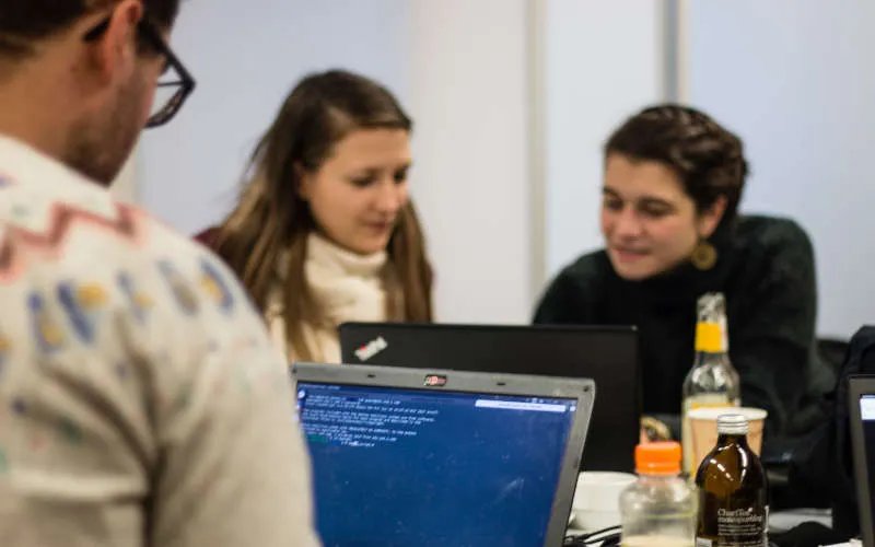 Drei Personen sitzen an ihren Laptops. Zwei von ihnen unterhalten sich.