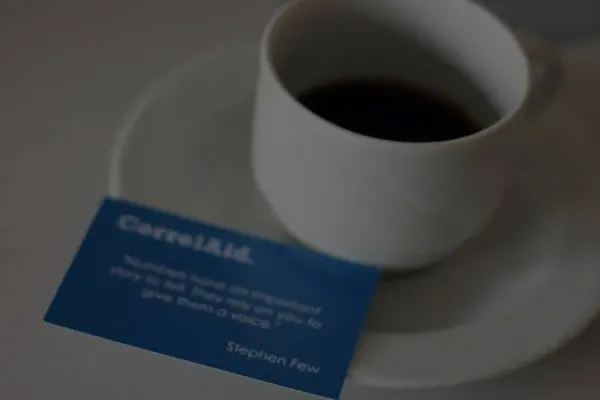 eine  halbvolle Kaffeetasse auf deren Untertasse eine Visitenkarte liegt. DIe Visitenkarte ist blau und hat die folgende Beschfiftung: CorrelAid.  Dann ein Zitat von Stephen Few, welches aber nicht richtig zu lesen ist. 
