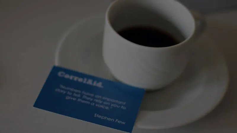 eine  halbvolle Kaffeetasse auf deren Untertasse eine Visitenkarte liegt. DIe Visitenkarte ist blau und hat die folgende Beschfiftung: CorrelAid.  Dann ein Zitat von Stephen Few, welches aber nicht richtig zu lesen ist. 